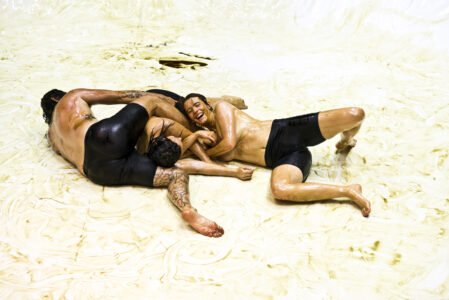 Three people in black short leggings and no top lie knotted together in a dynamic movement on a dirty white floor. Their skin is oily smeared. The face of a laughing person is turned towards the camera.
