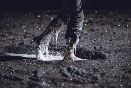 A dark floor is covered with black glittering rubble. A pair of feet, from which a white viscous substance drips, take a step in a small crater in the rubble, about 1 metre wide.