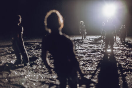 In a dark stage space, six people stand with their backs to the camera, facing a bright light. They sink to their knees in black glittering rubble and all lean slightly backwards.
