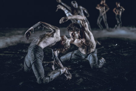 On a stage covered with glittering black rubble, two topless people dance on their knees, bending towards each other, one backwards, the other forwards. In the background, four blurred people dance upright.