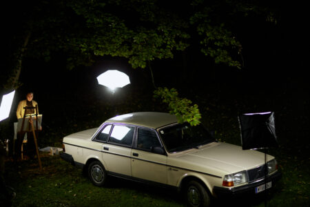 A white car is photographed in the dark under trees with a professional lighting setup.