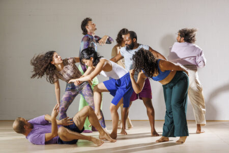 A group of dancers perform in a bright room in front of a white wall. They take different poses