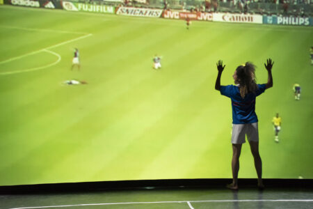 Vor einer großen Leinwand, auf die ein Fußballspiel projiziert wird, steht eine Frau in einem blauen Trikot und weißen Shorts und legt ihre Hände auf die grüne Rasenfläche.