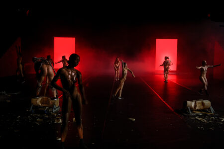 Eight naked performers stand separately in a large stage space in different poses, several have a kind of skeleton strapped on like a rucksack. Two projection screens in the background bathe everything in reddish light.