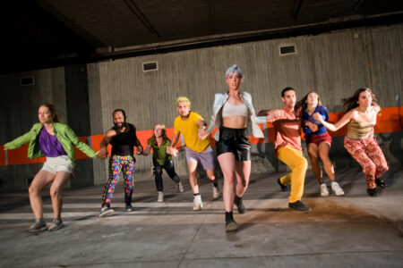 Eight diverse performers in colorful, simple costumes hold each other by the hands in a chain and walk towards the camera. The space is made of concrete and brightly lit.