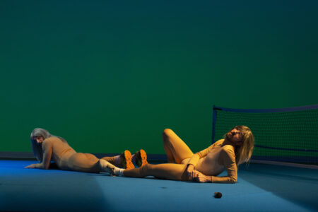 Two bearded performers in tight-fitting beige bodysuits and wigs lie on the floor, one on his stomach, the other propped up on an elbow. Their feet are touching, with a badminton net in the background.