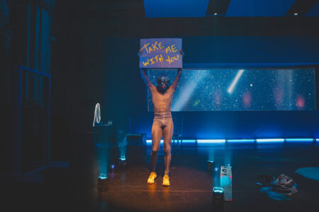 Raja Feather Kelly wears a shimmering, semi-transparent bodysuit and bright yellow trainers. He holds up a poster with the inscription Take me with you. In the background is a projection of the Milky Way.