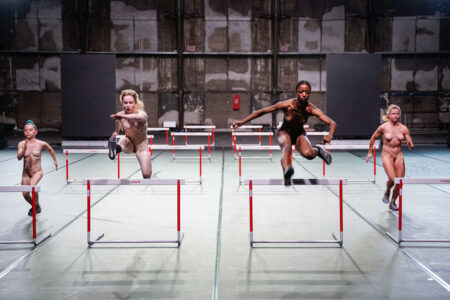 In a factory-like stage space with a white floor, four naked performers run over hurdles towards the audience, the two in the middle are jumping.
