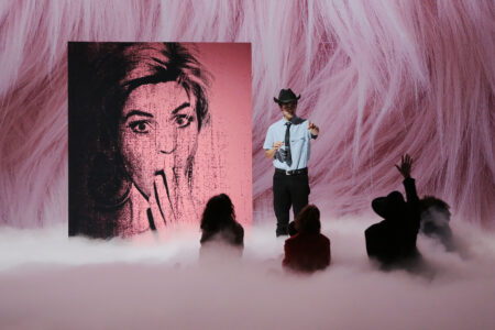 Several actors are sitting around a standing actor in a blue shirt and cowboy hat in a cloud of dry ice, in the background a large portrait of a woman and fluffy pink flokati carpet.
