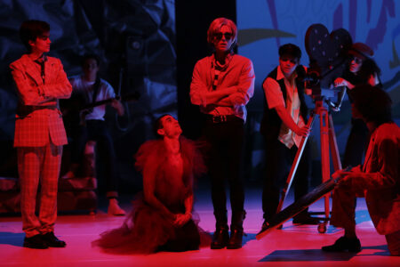 In the center of the photo stands an Andy Warhol look-alike with his mouth slightly open, around him some actors are facing him. One kneels at his feet, others operate a large film camera.