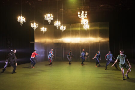Eight dancers position themselves in a large semicircle, their legs are slightly bent and they stretch their right arm into the center of the circle. The stage space is lit in pale green, nine chandeliers hang from the ceiling.
