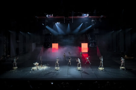 In the predominantly black stage space, there is again a lot going on: cars hanging from the ceiling in the background, two long staircases to the sides, a large number of naked performers spread out on the stage, working on short tree trunks with chainsa