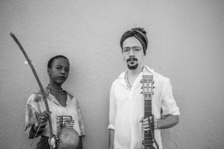 Black and white photo of dumama and kechou, she in short blouse with writing on it, short black hair and tight necklace, he in white shirt, short beard and cloth ribbon in hair. Both are holding their instruments.