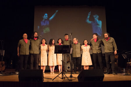 A 10-piece band stands smiling between microphones and monitors arm in arm on a stage. The women wear beige shirt-dresses, the men dark trousers and camouflage green shirts. They all wear red neckerchiefs.
