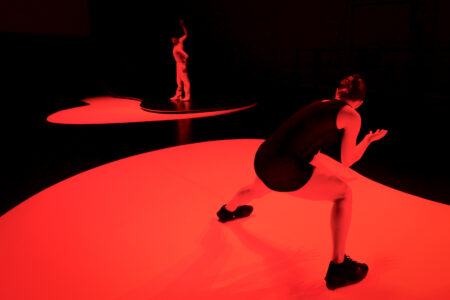In a very dark, red-lit stage space, two dancers stand on round, curved stage elements. The one in front is in a dynamic crouch, the one behind is standing straight with arms stretched upwards.