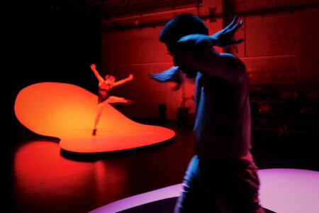 In a dark, reddishly lit stage space, two dancers stand on rounded, curved stage elements. The one in the back throws her arms and one leg in the air, the one in front swings her hands around her head.