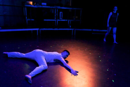 A man in white clothing lies stretched out on a dark stage floor onto which an orange light spot falls. He reaches with one hand into the centre of the circle of light. In the background, a woman walks along the edge of the stage.