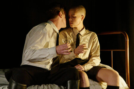 Two people in white shirts, dark ties sit on a bed and almost kiss. Both are holding filled white wine glasses.