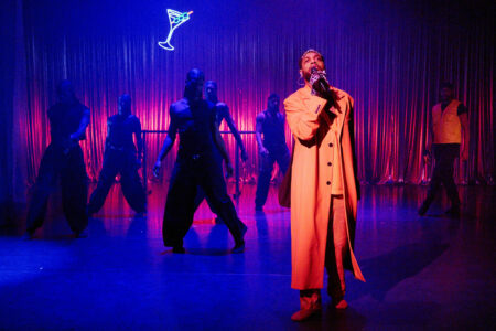 Vor einem blau beleuchteten Vorhang steht ein Schwarzer Mann in einem eleganten orangenem Mantel und singt in ein Mikrofon. Hinter ihm stehen fünf Männer in dunkler Kleidung breitbeinig und rechts ein kommt ein Mann in orangener Weste ins Bild gelaufen.