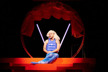 A person in a blue life jacket and a blue net wrapped around their legs sits in front of a circle with a red curtain and two white neon lights. The person has a scarf on his head, their hands gently placed in their lap and speaks.
