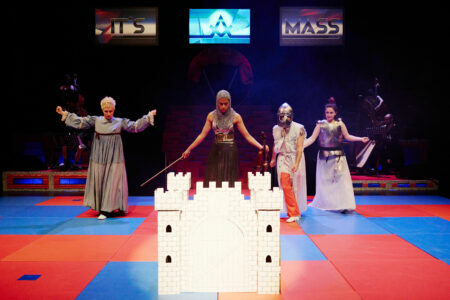 On a red and blue tiled stage floor, four people in medieval costumes stare at a white castle about 50cm high. One person in a knight's costume holds a sword and looks particularly intense. In the background are the words IT'S A MASS.