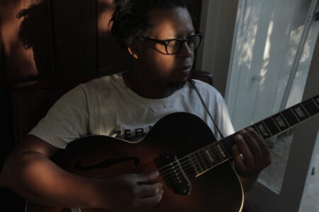 A Black man with glasses and a white T-shirt sits at a windowsill and plays an acoustic guitar. Small rays of sunlight are on his face and on the wall behind him.