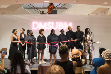 9 Schwarze Menschen, alle bis auf eine Frau am Mikrofon, schwarz gekleidet, stehen auf einer Bühne hinter einem roten Band, das zur Eröffnung der Ausstellung durchgeschnitten werden soll.