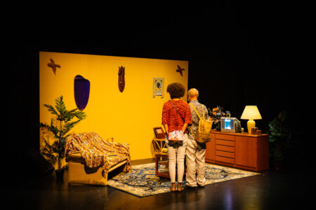 Zwei Menschen stehen in der Installation und betrachten ein "Wohnzimmer" das aus einer gelben Wand besteht, links steht ein Sofa, in der Mitte ein Tisch und rechts ein Sidebord mit Fernseher, Lampe, Pflanzen und anderen Gegenständen.
