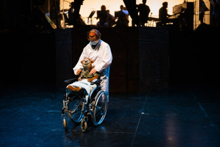 A bear puppet sits in a wheelchair in the middle of the stage. The wheelchair is pushed by the puppeteer, who is wearing a white coat. The musicians sit on a stage in the background.