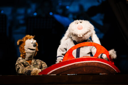 Zwei Puppen, links ein Bär und rechts ein Hase sind auf der dunklen Bühne und vor dem Hasen ist ein rotes Lenkrad.