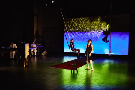 Zwei Performerinnen (in der Mitte) und zwei Musiker (links) sind auf einer dunklen Bühne und im Hintergrund ist eine Videoprojektion.