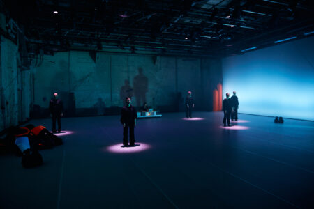 Fünf Performer*innen stehen mit viel Abstand verteilt in einem blau beleuchteten Bühnenraum. Sie werden einzeln von vertikalen, lila Lichtspots angestrahlt.