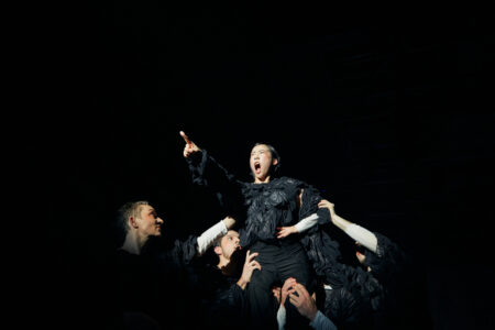 A performer in a black costume is lifted up by four others. She has her mouth wide open, her eyes angry. She points her outstretched index finger upwards and follows it with her gaze.