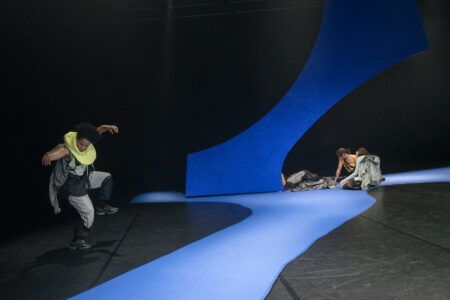 In einem dunklen Bühnenraum mit blauen, zackigen Elementen auf dem Boden und der Wand steht eine Person im Vordergrund und tanzt mit erhobenen Armen und einem Bein in der Luft. Im Hintergrund sitzen drei Personen im Schatten eines blauen Elementes.