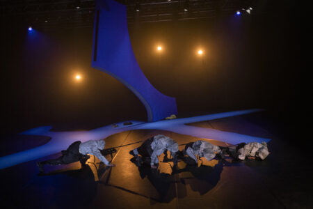 Four people in silver and black costumes with hoods crawl on all fours in a row on the floor in a dark stage space. A blue-lit bent column stands in the background and yellow spotlights shine.
