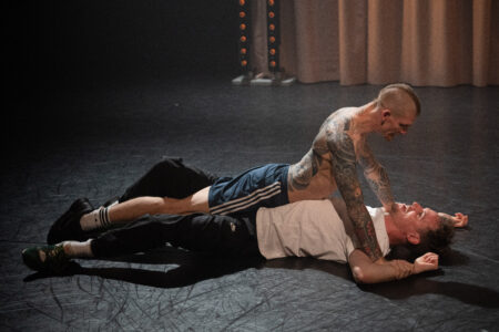 A heavily tattooed white man, topless and wearing Adidas shorts, lies on top of a white man in black trousers and a white Tshirt, pressing the latter's arms to the floor next to his head. They gaze intensely into each other's eyes, their legs entangled