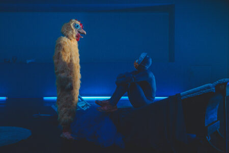 Raja Feather Kelly sits on a slightly elevated stage element with his knees drawn to his chest, looking up at a performer in a yellow chicken costume, both in profile, all bathed in blue light.