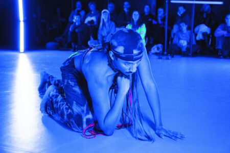 A man of Colour with a moustache kneels on a blue-lit stage floor and supports his chin with his fist. He wears batiked corduroy trousers and a durag with the same pattern. Behind him, the audience sits on the floor and on small stools.
