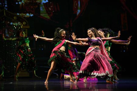 A Bollywood dance scene on a dark stage. Four dancers in pink-green costumes, golden jewelry and long black hair stand wide-legged and with arms stretched out to the sides, looking happily at each other.