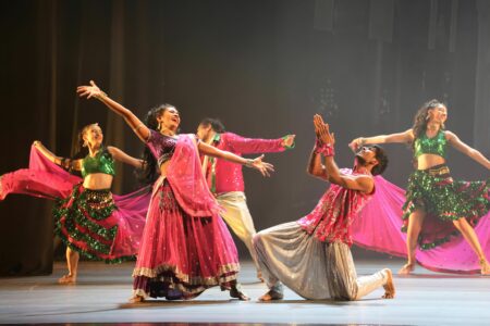 A Bollywood dance scene. Five dancers in pink and green costumes and golden jewellery are standing on a stage, smiling happily with their arms stretched out to the sides. One at the front kneels on one leg and worships one of the female dancers.