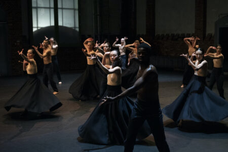 12 dancers stand loosely distributed and moving synchronously in a dark room. They are topless and wear either very wide, dark blue skirts made of sturdy fabric or dark blue trousers. They each stretch one arm forward in a demanding manner.