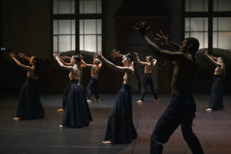 9 dancers are loosely distributed in a dark room with large windows. They are topless and wear either very wide, dark blue skirts made of sturdy fabric or dark blue trousers. They are turned to the side and stretch their arms upwards.