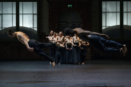 In a room with large windows, about 8 topless people with long dark skirts stand against the wall and clench their fists. In the foreground, two people are jumping almost horizontally about one metre above the floor with their arms stretched out backwards
