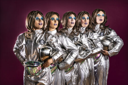 Five women with 60s-style wigs, bright blue eye shadow and dark purple lipstick stand close together in a row. They wear shiny silver overalls and hold metal pasta strainers under their arms. They look to the side, startled.