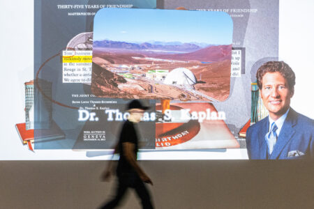 A person in a black outfit walks through a projection. It shows a collage of two bookcovers overlayed by a photo of a research facility and a portrait of a man, smiling. A person in a black outfit crosses through the projection.