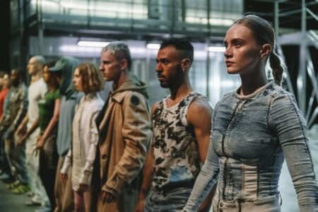 Ten people in different, artistic costumes reminiscent of streetwear stand in a row. The person in front on the right edge of the picture is sharp, the people further back become increasingly blurred. They all stoically look ahead.