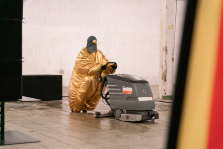 Eine Person in einem aufgeblasenen goldenen Bodysuit trägt eine Taubenmaske auf dem Kopf und schiebt eine Poliermaschine über den Boden. Am Bildrand ragt eine Deutschlandfahne hervor.
