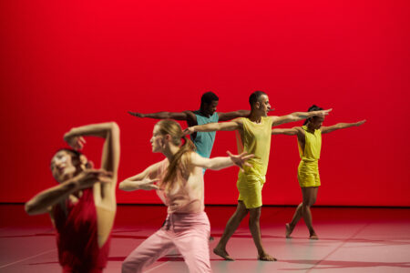 Fünf Tänzer*innen in einfarbigen Kostümen in jeweils Gelb, Rosa, Rot und Hellblau, stehen mit ausgestreckten Armen in zwei Gruppen vor einer knallroten Wand.