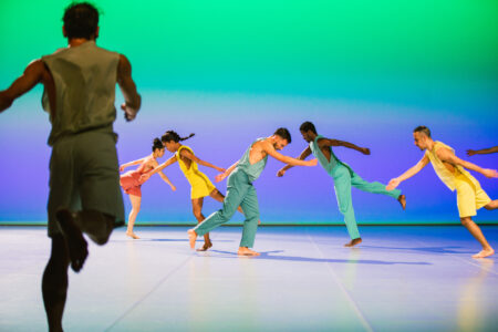 5 Tänzer*innen in gelben, türkisen, roten Kostümen, laufen vor einem blau-grünen Hintergrund in großen Fallschritten. Im Vordergrund läuft eine Person auf die Gruppe zu.