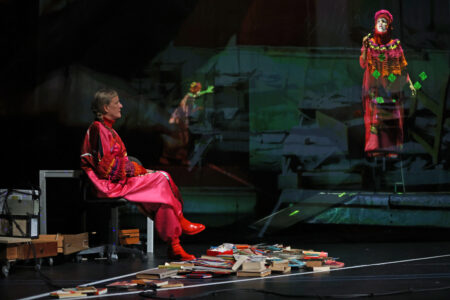 The photo shows a performer in a red costume sitting on an office chair in front of a table in the middle of a pile of books in the foreground. In the background, two other performers can be seen exploring the stage space with torches.